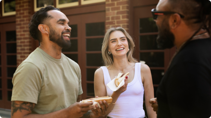 Three people smiling at eacch other whilst eating a hotdog